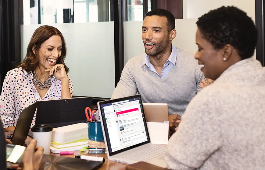 Three people sit at a table with laptops and notebooks, smiling and talking in a modern office setting. One woman holds a coffee cup, and everyone appears engaged while discussing ideas using Office 2019 Professional Plus during the meeting.