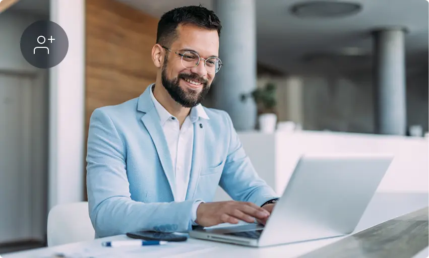 A smiling man with a beard and glasses, wearing a light blue blazer, sits at a desk working on a laptop in a bright, modern office. A user profile icon with a plus sign is visible in the corner.