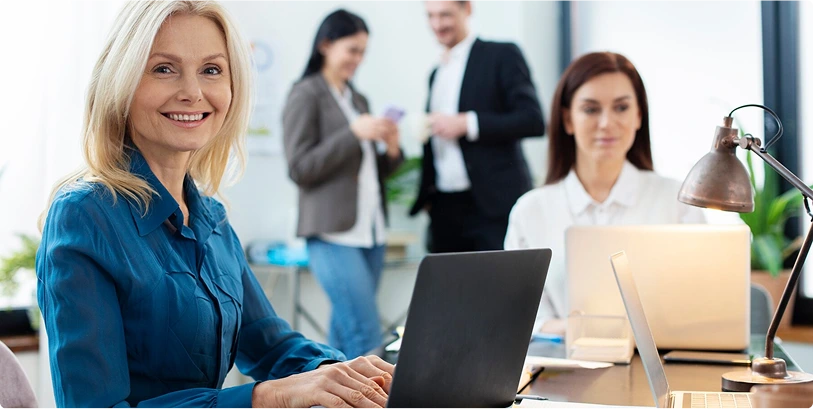 A woman in a blue blouse smiles at the camera while working on a laptop at an office desk. Another woman works nearby, and two people are talking in the background. The setting is bright and professional.