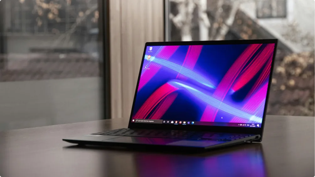 A sleek, thin laptop with a vibrant screen displaying abstract blue and pink lines sits on a wooden table beside a window with blurred trees and buildings in the background.