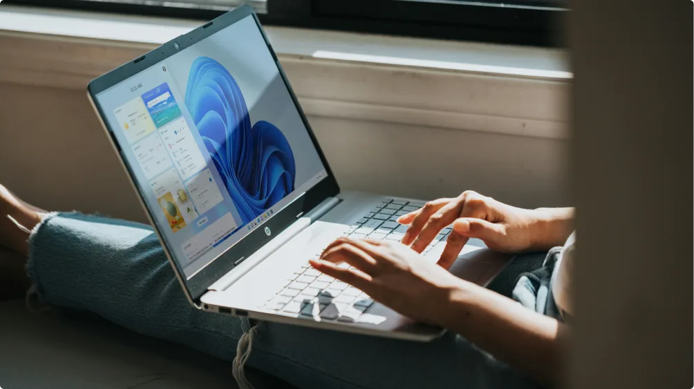 Person sitting on the floor, typing on an HP laptop with Windows 11 interface on the screen, sunlight streaming through a nearby window.