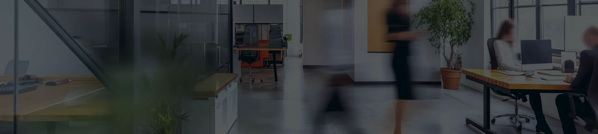 Modern office space with desks, computers, a large potted plant, and glass walls. Two people are sitting at desks working, while two blurred figures walk through the scene, suggesting motion.