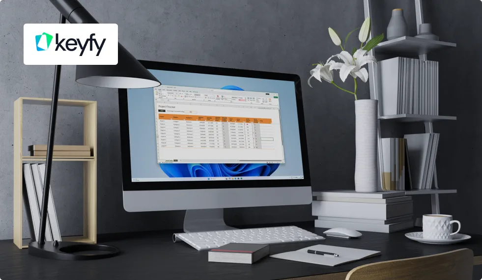 A modern desk setup with an iMac displaying a spreadsheet, a keyboard, lamp, books, a white coffee cup, and a vase with white flowers. The Keyfy logo is in the top left corner of the image.