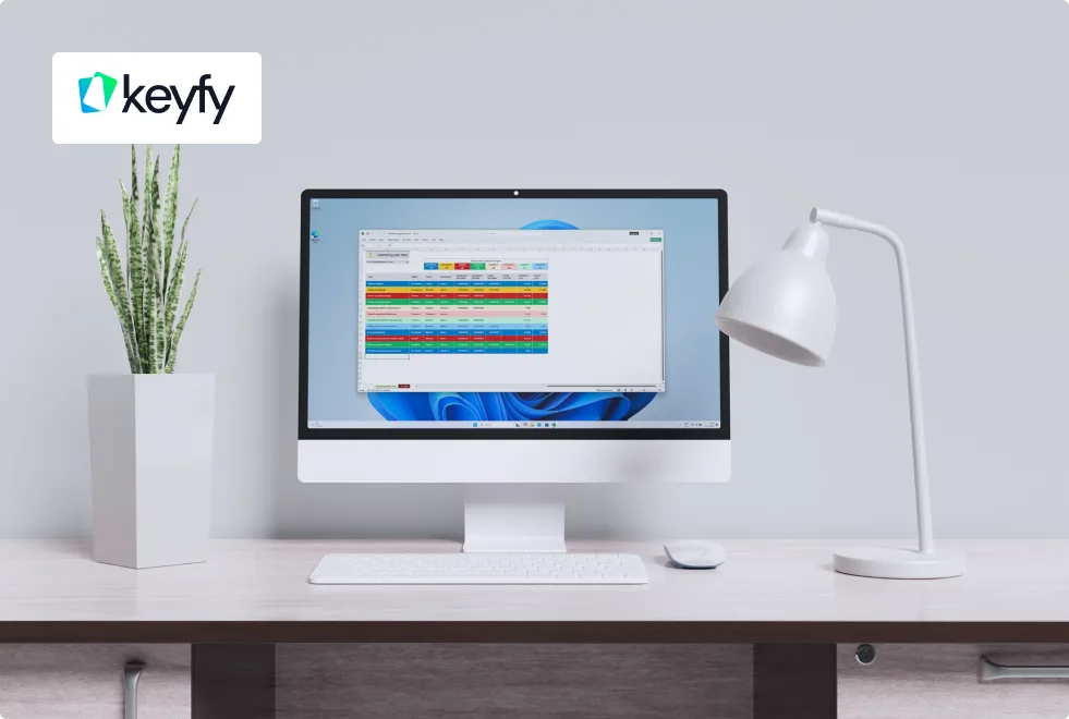 A modern desk with a white computer, keyboard, mouse, plant, and lamp. The screen displays a colorful spreadsheet, and the keyfy logo appears in the top left corner.