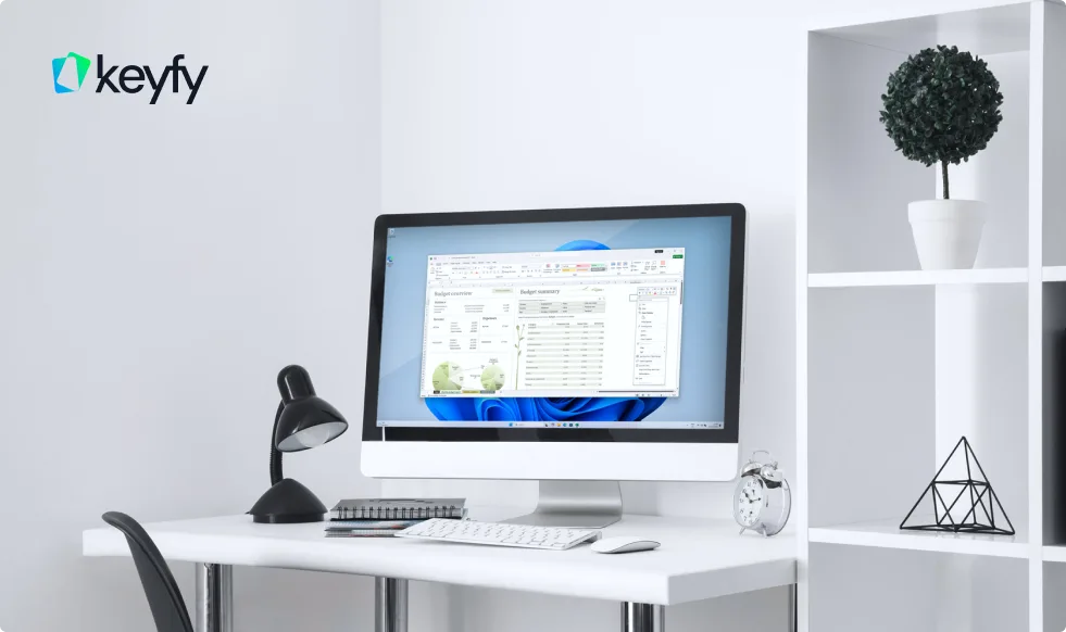 A clean white desk with a desktop computer displaying a spreadsheet, a black lamp, a notebook, an alarm clock, and a shelf with a small plant and decorative items. The Keyfy logo appears in the top left corner.